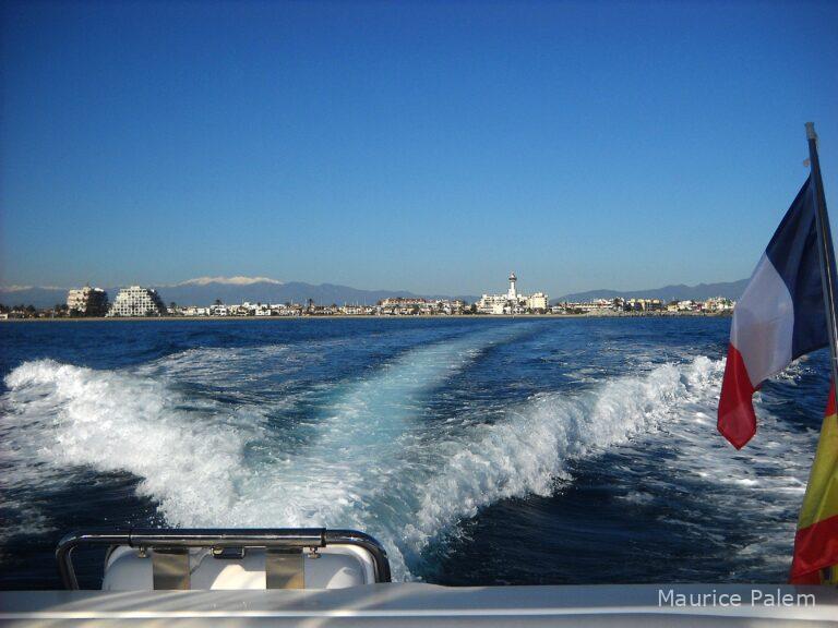 Vue de la baie d’Empuriabrava depuis l’arrière d’un bateau sous pavillon français, avec la ville et les montagnes en arrière‑plan.